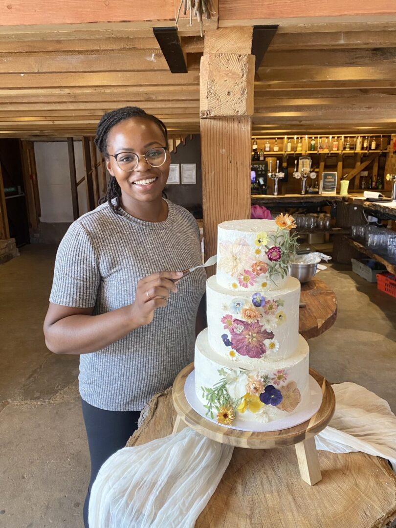 Person smiling with glasses and a striped shirt stands beside a three-tiered cake decorated with colorful edible flowers, holding a spatula. Rustic wooden beams are visible in the background.